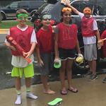 Four students with goggles and sponges wash a car