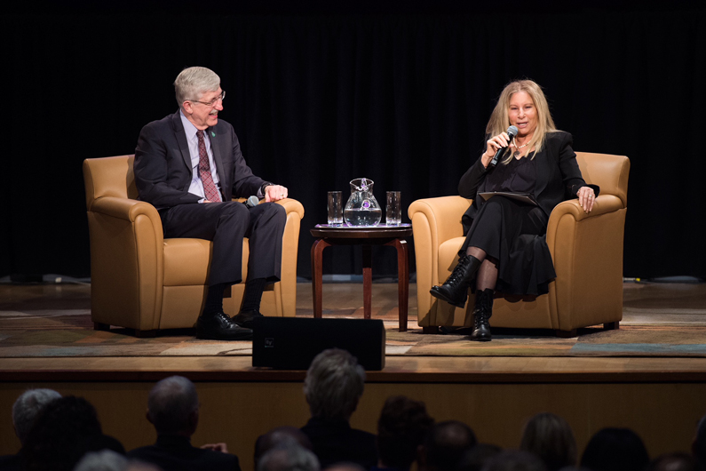 Dr. Francis Collins, director of the NIH sties in a chair to the left. Barbra Streisand holds a microphone and sits in a chair to the right – speaks to the audience.