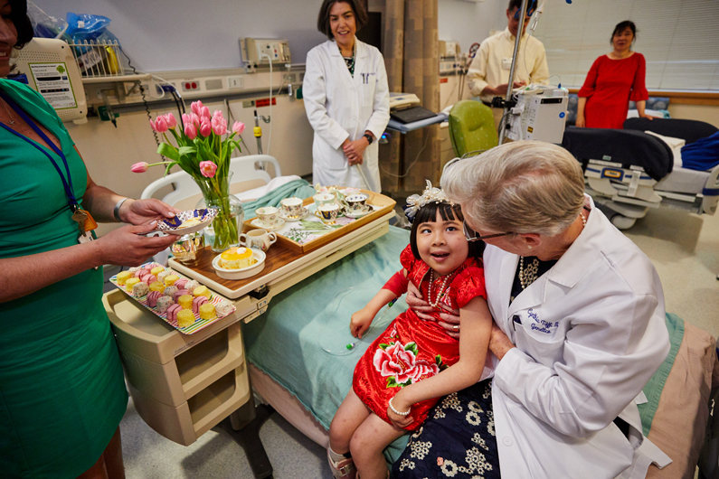 Pediatric patient sits on the bed with her care provider, as a tea set, tea cakes and flowers are on display in celebration of her gene therapy